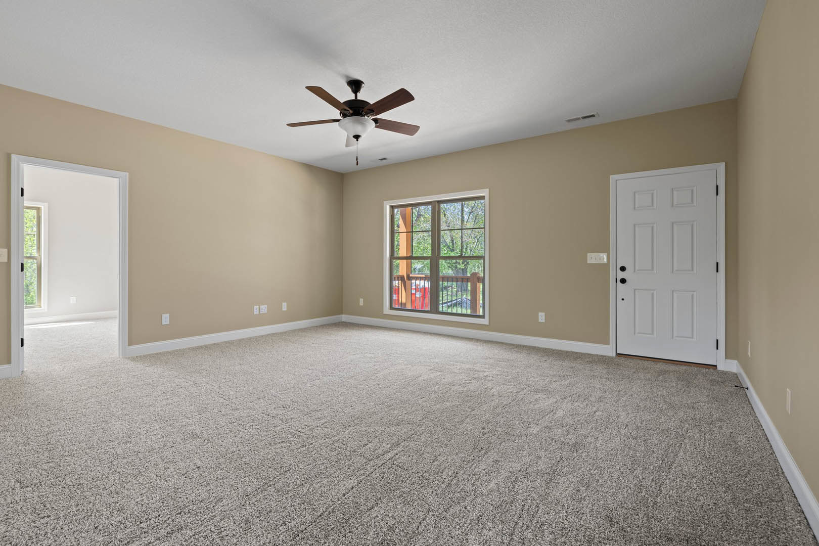 Ceiling fan with light fixture above carpeted floor, white walls, white door with black knobs, window overlooking trees and railing