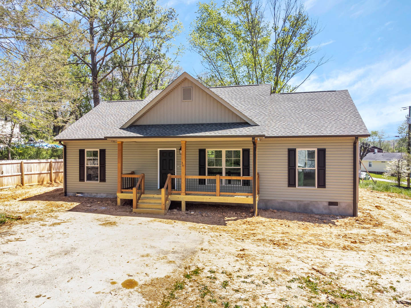 Two-story house with white siding, black shutters, and black front door, front porch with stairs, concrete driveway, wooden fence with posts, mature trees and lawn in foreground