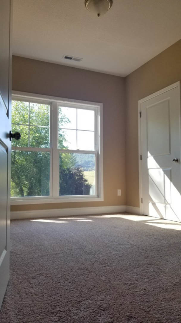 Bedroom with beige carpet, white walls, large window overlooking trees, open white door with silver handle, partial view of toilet in adjacent bathroom