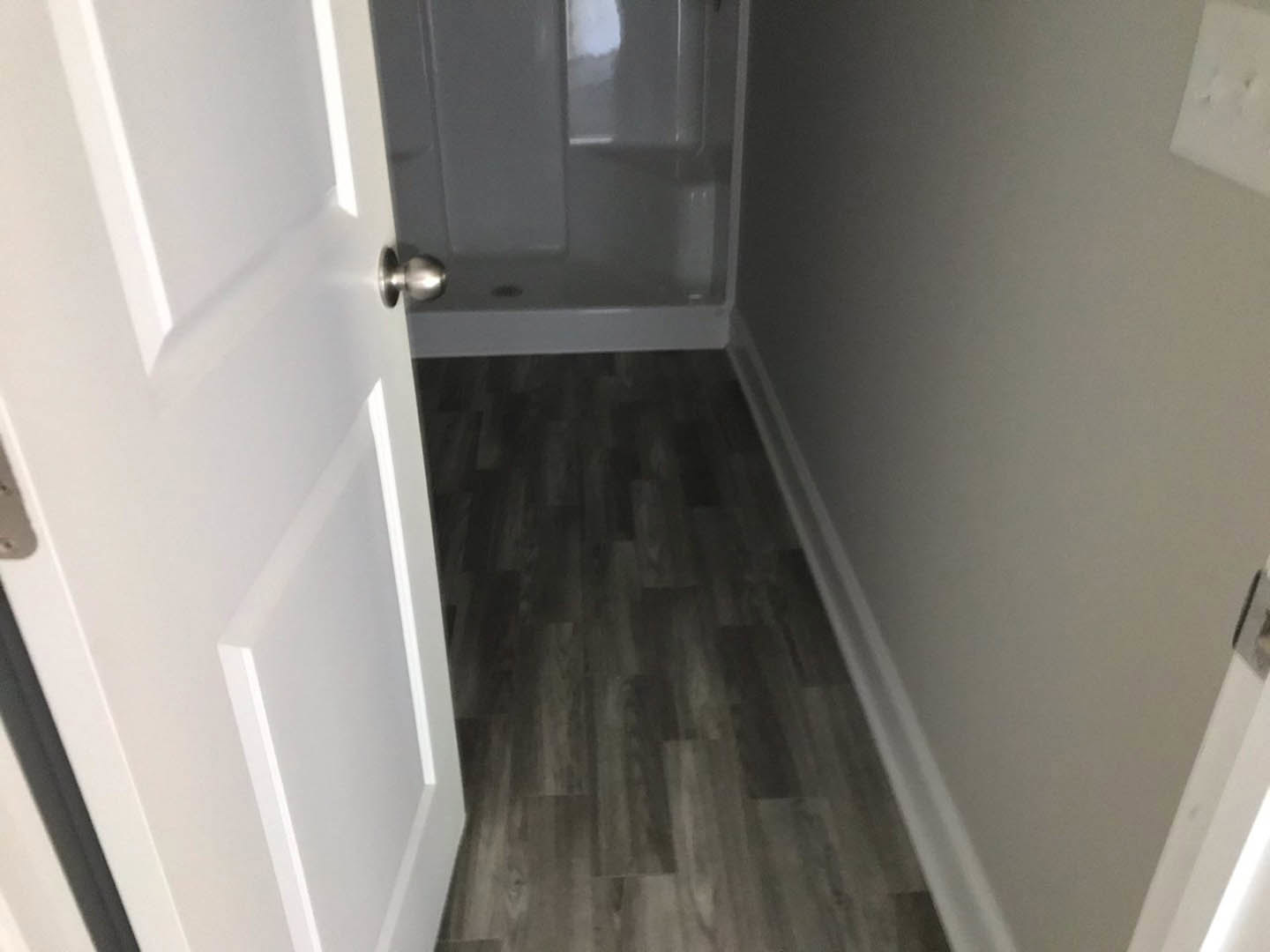White bathroom door with silver knob, dark wood flooring with white trim, partial view of shower stall and white wall surface
