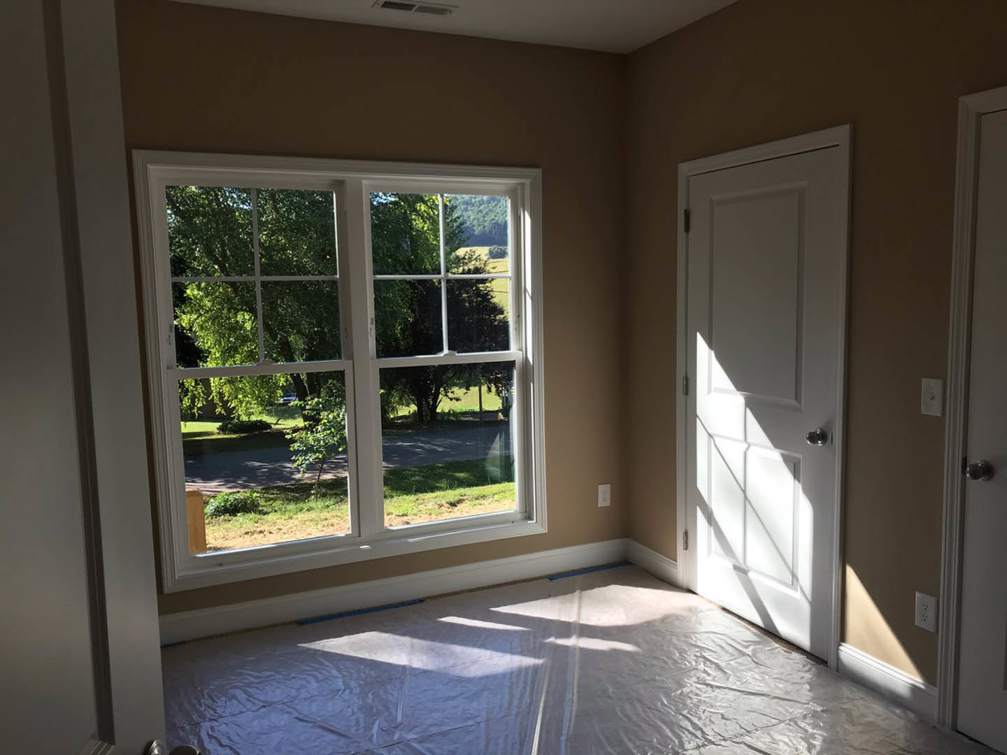 White-walled room with a white door featuring a silver knob, large window showing trees outside, white sheet spread on hardwood floor
