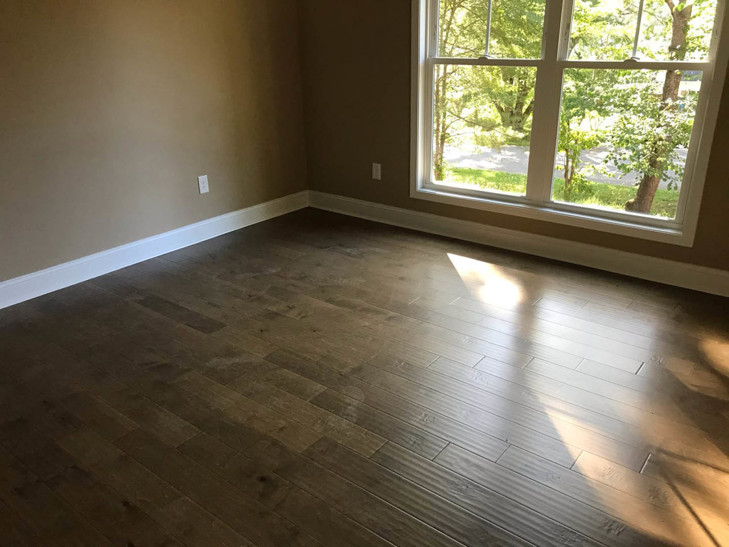 Wood flooring in a bright room with a large window overlooking leafy trees, white walls, and natural daylight.