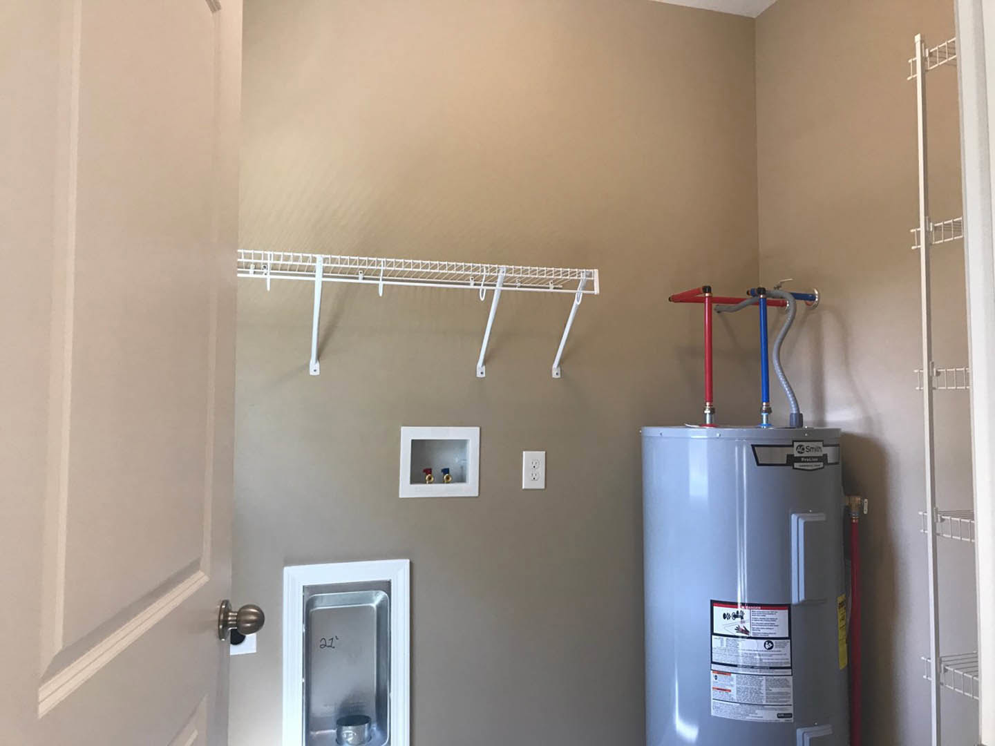 White tankless water heater mounted on plaster wall, red and blue pipes connected below, rectangular metal tray with number, small window on white box, indoor bathroom space.