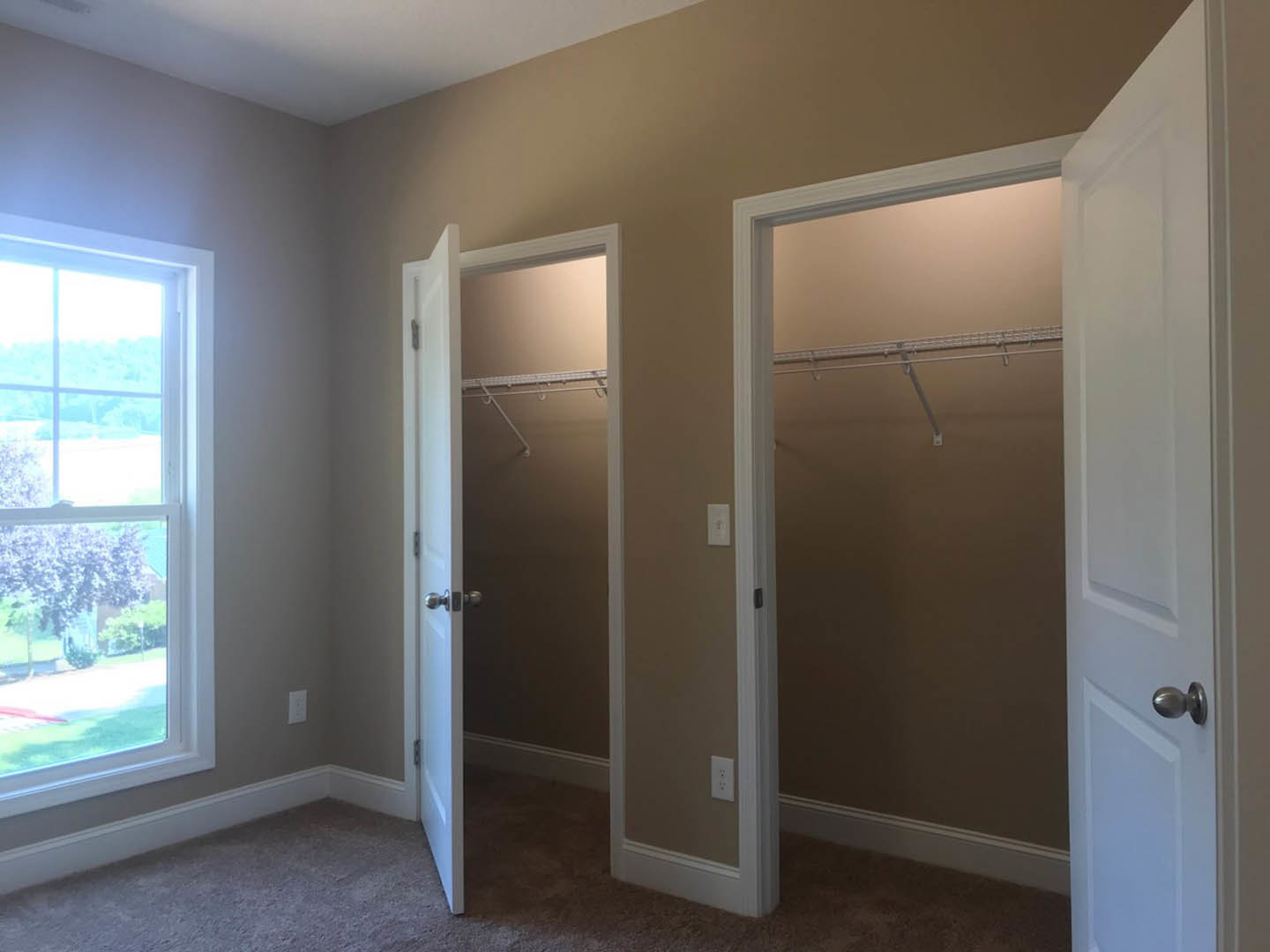 Walk-in closet with two white doors open, shelving and hanging rods visible, window overlooking green trees and grass, brushed metal door handle, light wood flooring