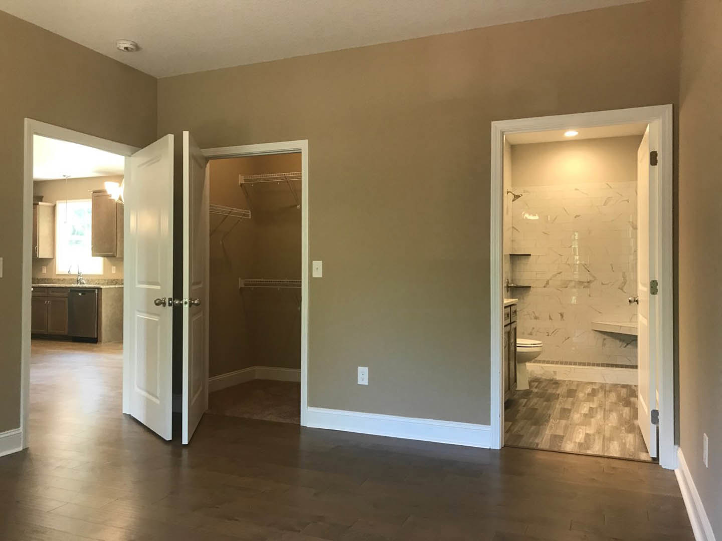Bathroom with wood-look laminate flooring, white walls and trim, open door revealing a walk-in shower, toilet, sink, and illuminated closet.
