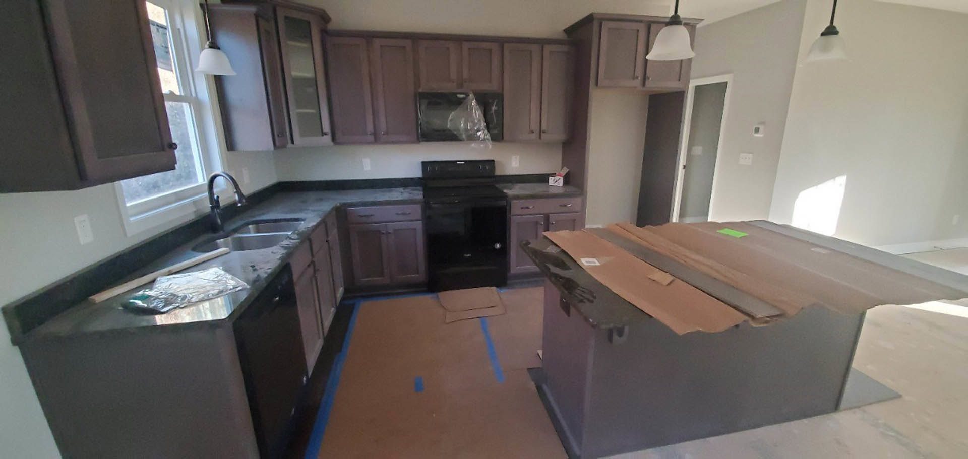 Kitchen with dark wood cabinets, black stove, silver cabinet knobs, cardboard box and plastic bag on table, white rectangular object on countertop, blue tape on floor