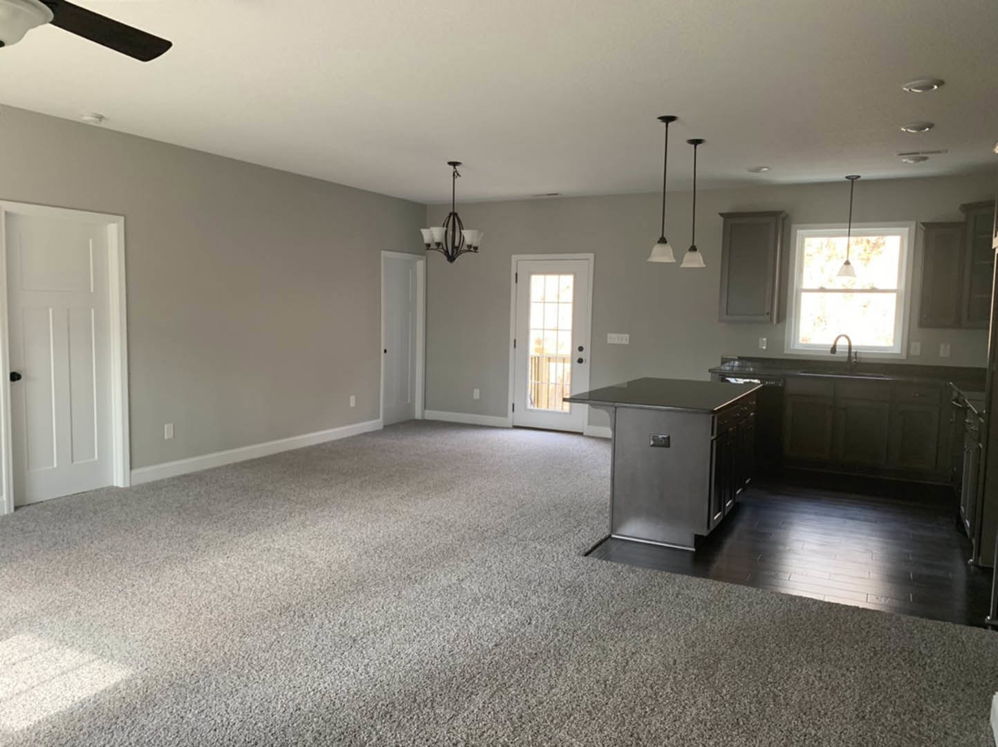 Open kitchen and living room with beige carpet, black countertop kitchen island, white cabinetry, white door with window and black knob, ceiling light fixture, neutral walls and