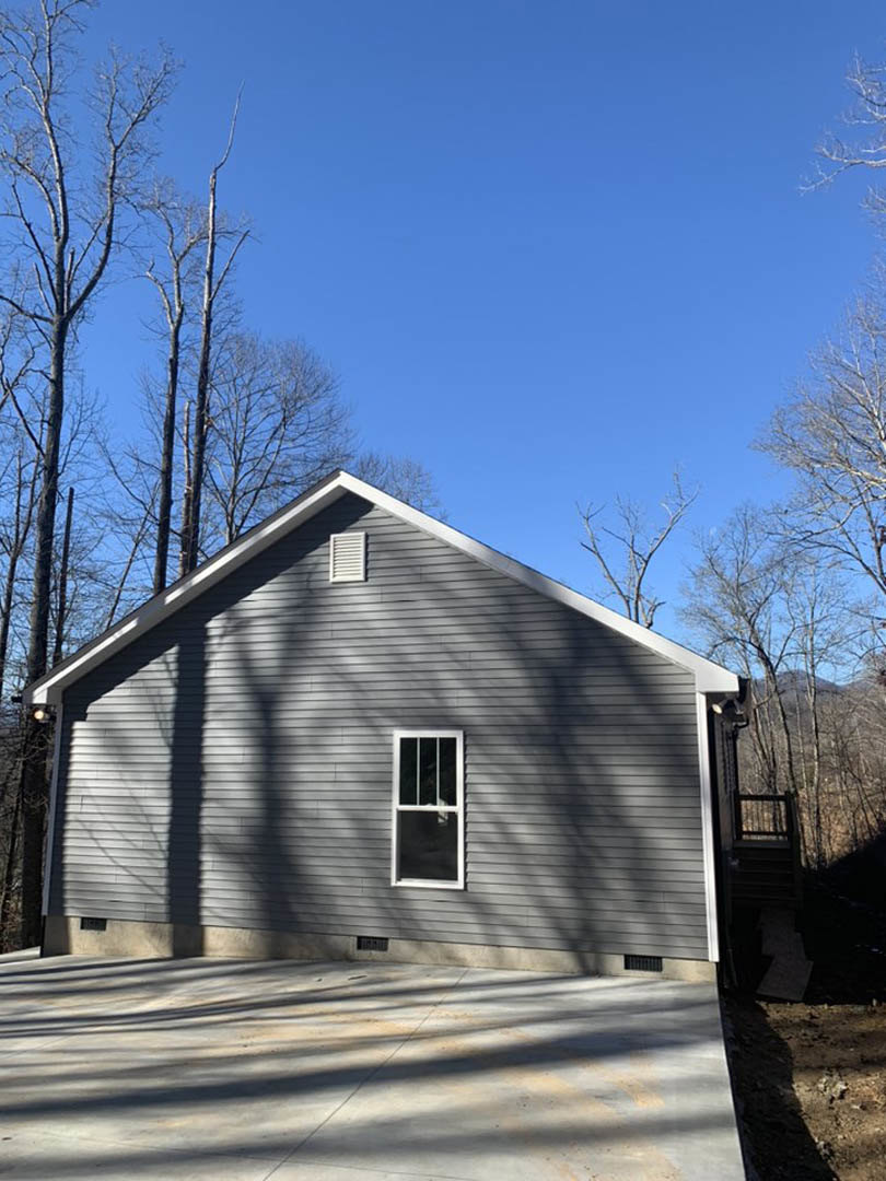 Modern house with white-framed windows, gray siding, and pitched roof, surrounded by mature trees, concrete walkway, and wooden fence in foreground