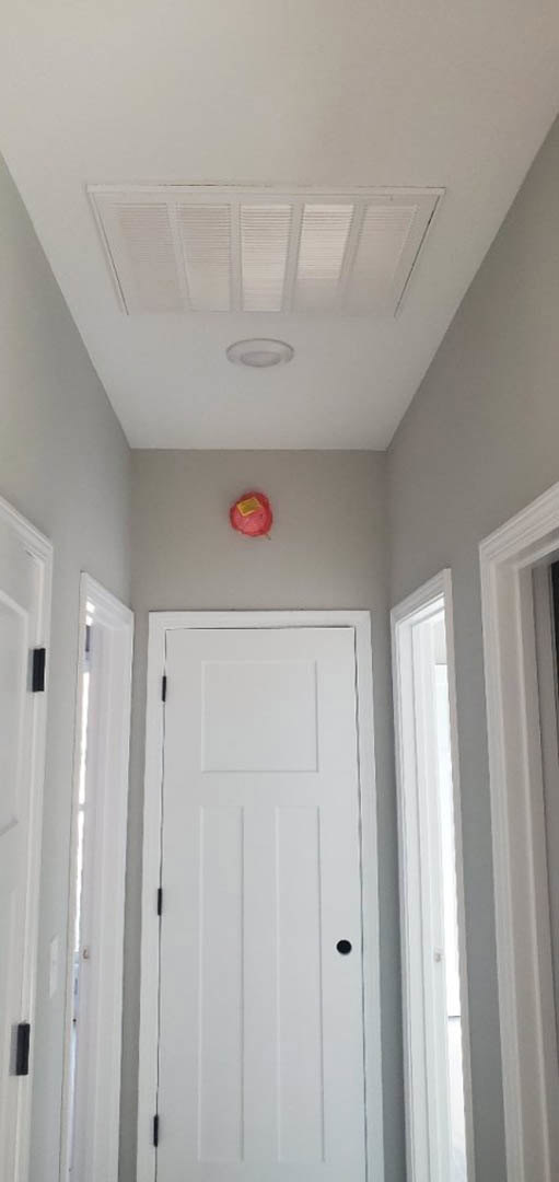 White paneled door with black handle set in white door frame, white ceiling with air vent, red wall-mounted object, pink item topped with yellow square.