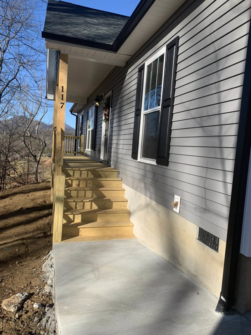 Wooden stairs lead to a covered porch on a house with light siding, large windows, and tree branches visible against a blue sky.