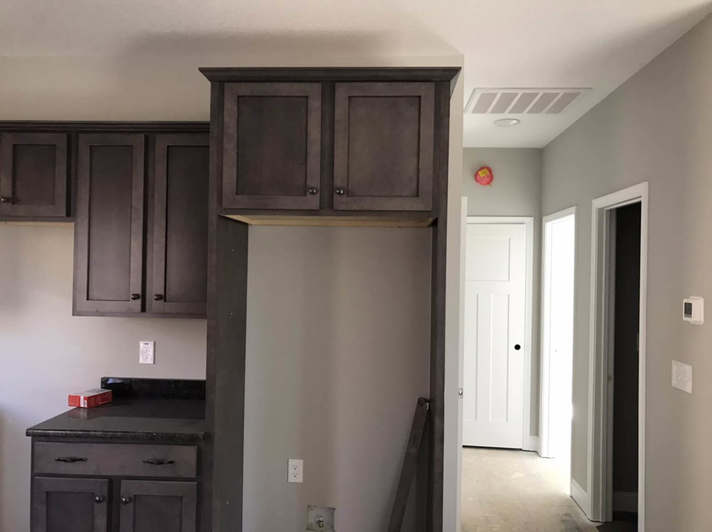 Dark wood kitchen cabinets, black countertop with a red storage box, white door featuring a black circular accent, close-up of cabinetry, white device with square screen on the