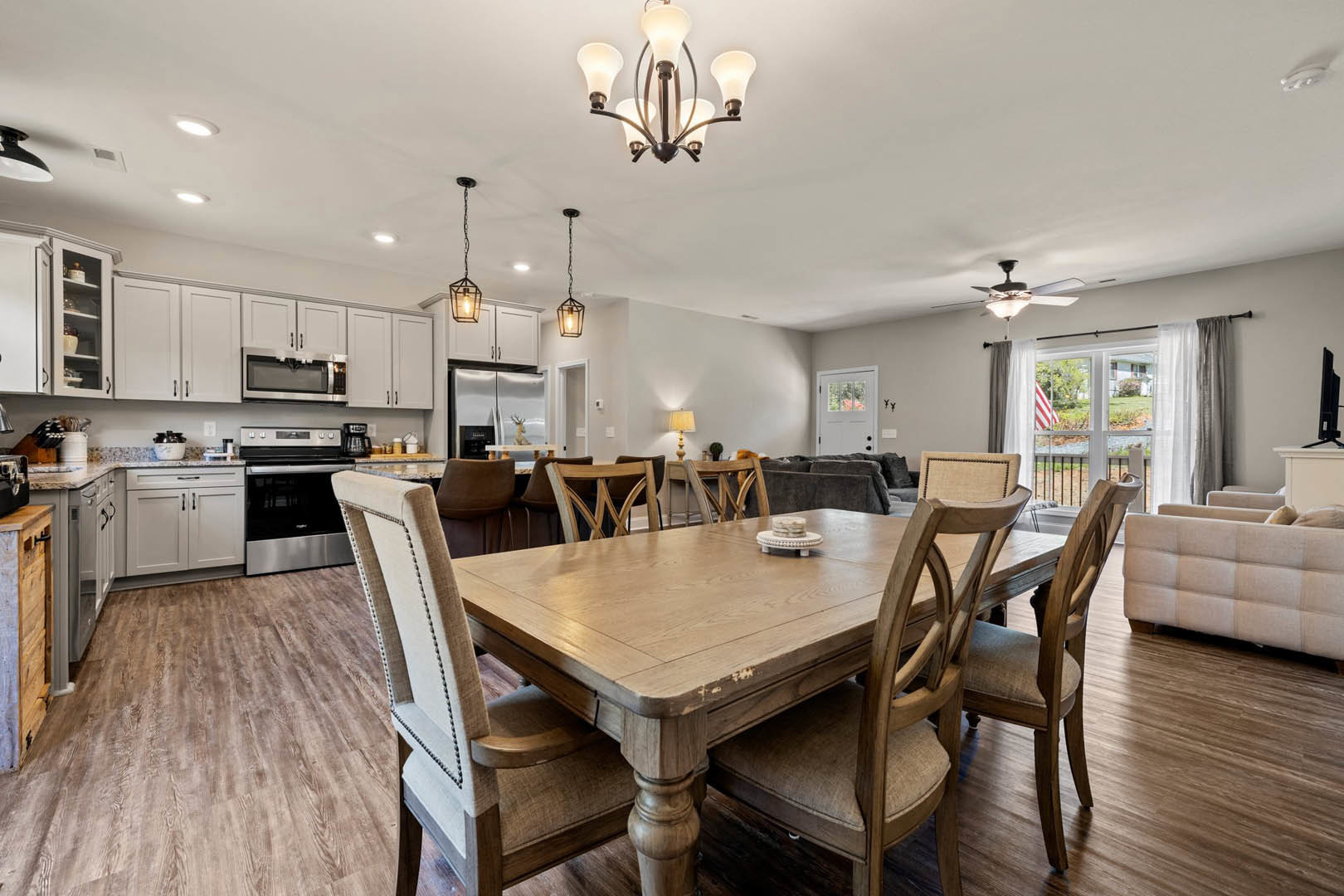 Wood dining table with upholstered chairs set beside a kitchen featuring white cabinetry, stainless steel stove, and five-light chandelier overhead.