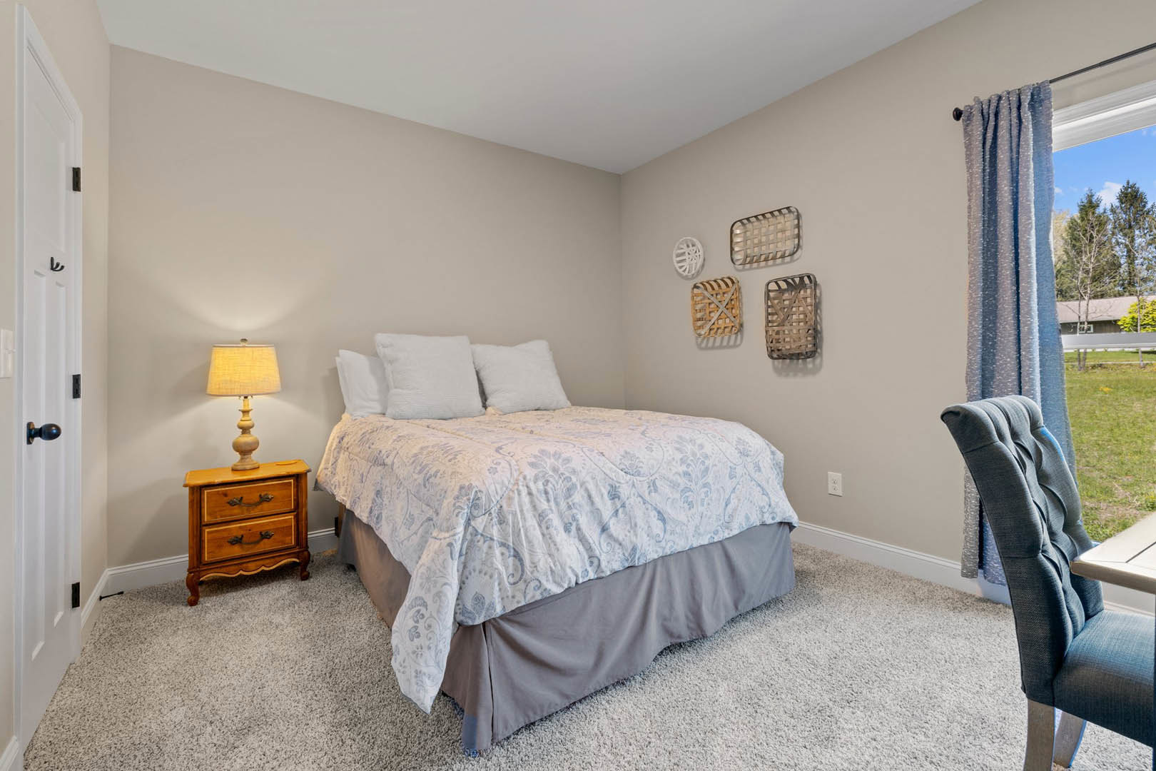 Bedroom with a bed featuring white pillows and a gray blanket, wooden dresser topped with a lamp, chair beside a small table, and decorative white circular wall art.