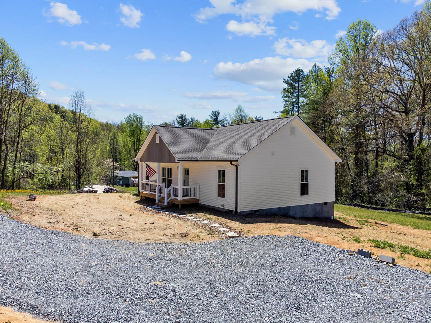White house with covered front porch, gravel driveway, American flag mounted near entry, blue sky with scattered clouds, mature trees and lawn surrounding exterior