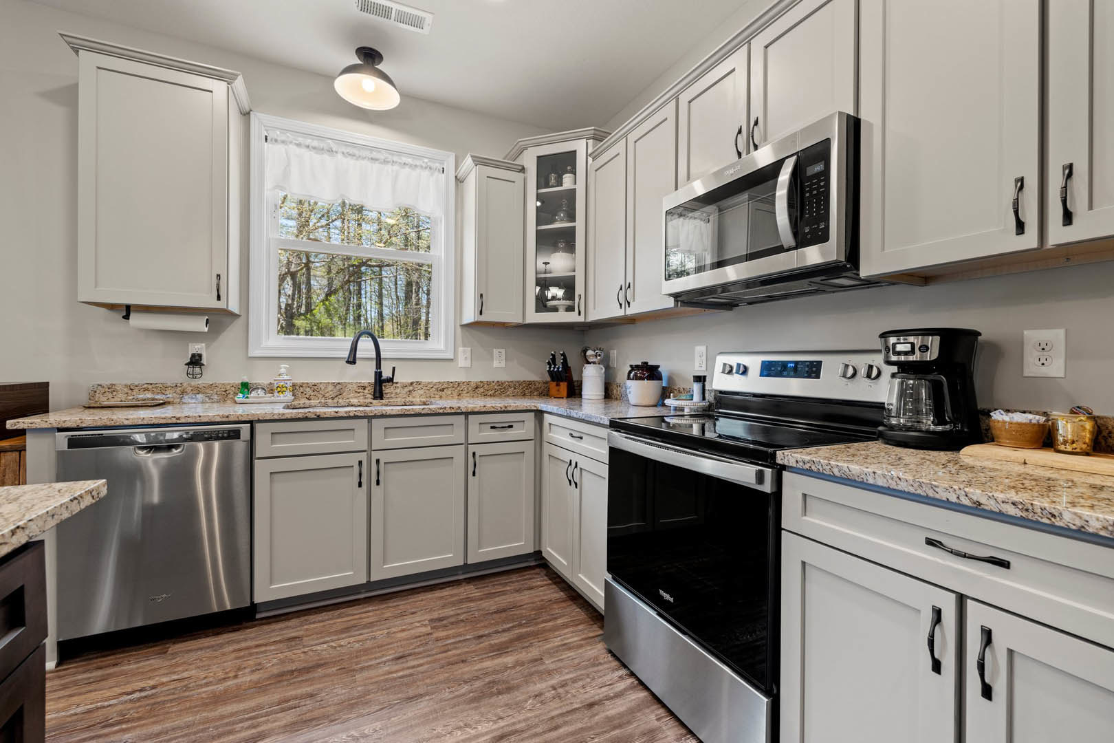 White kitchen cabinets, black stove and microwave, stainless steel dishwasher, light fixture on ceiling, window with white curtain and view of trees, light countertops and sink.