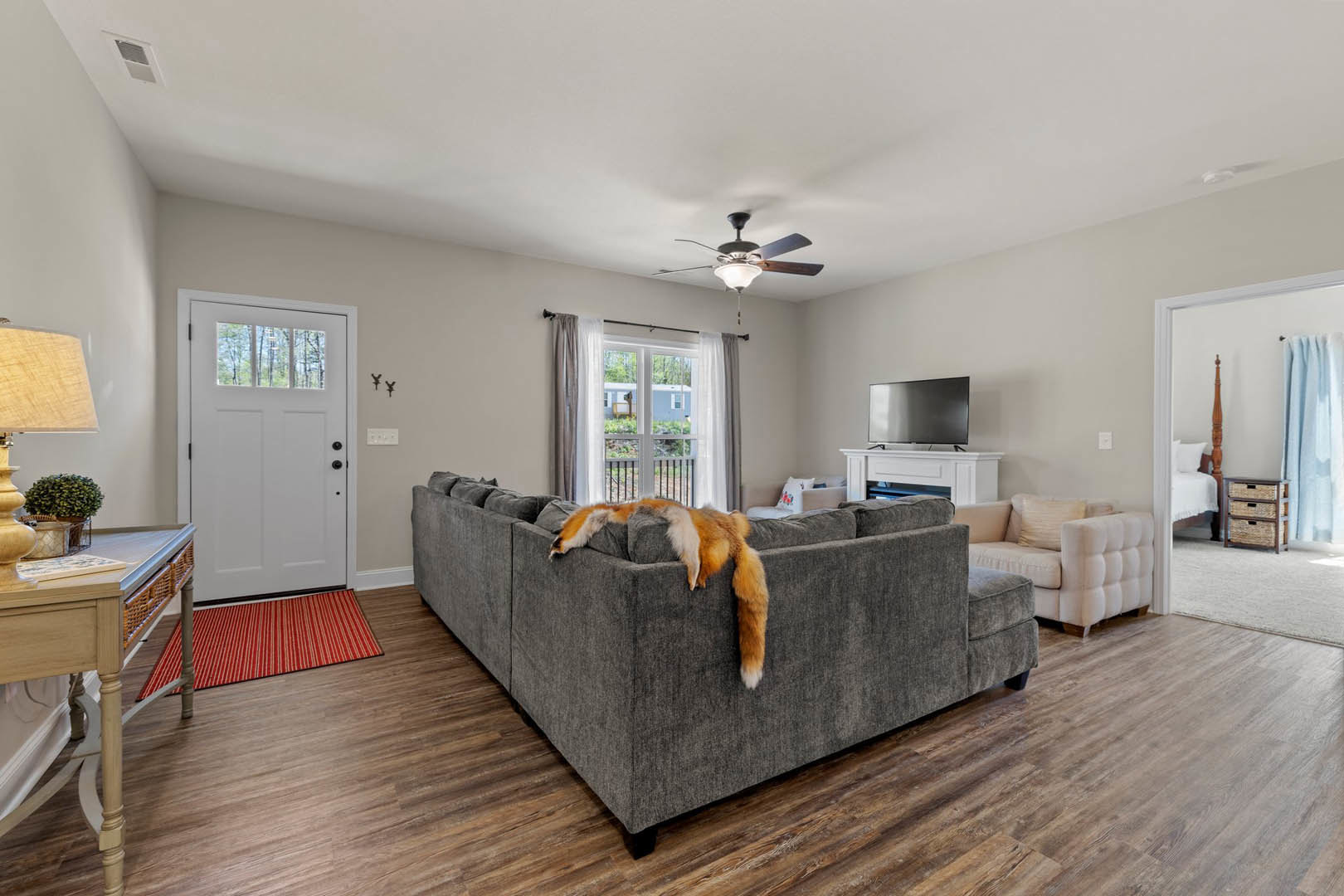 Living room with a gray couch, ceiling fan, red and white striped rug, white door with glass panes, window with curtains and railing, laminate wood flooring, and a person standing
