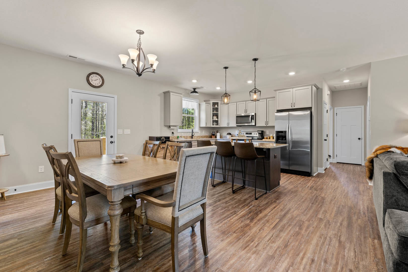 Open kitchen and dining area with wood table and chairs, stainless steel refrigerator with black door, white cabinetry, wall clock, white door with black knob, visible ceiling
