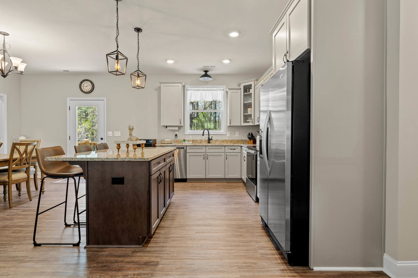 Kitchen with black refrigerator, wood flooring, white cabinetry, stainless steel sink, wall clock, window with white curtains, and wooden seat chair