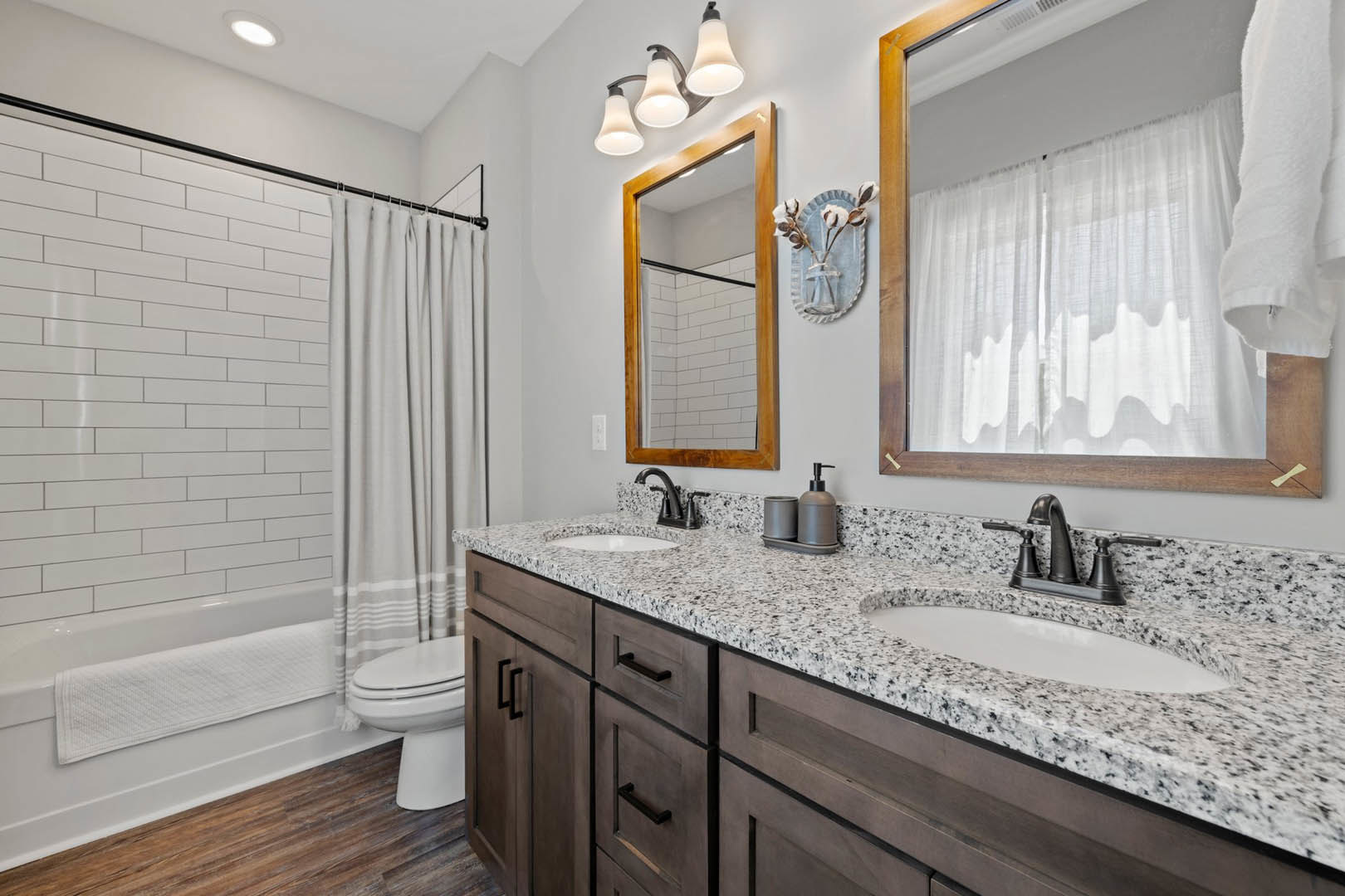 Bathroom with marble countertops, white tile walls, shower curtain, large mirror, three-light fixture, and a vase of cotton flowers on the vanity