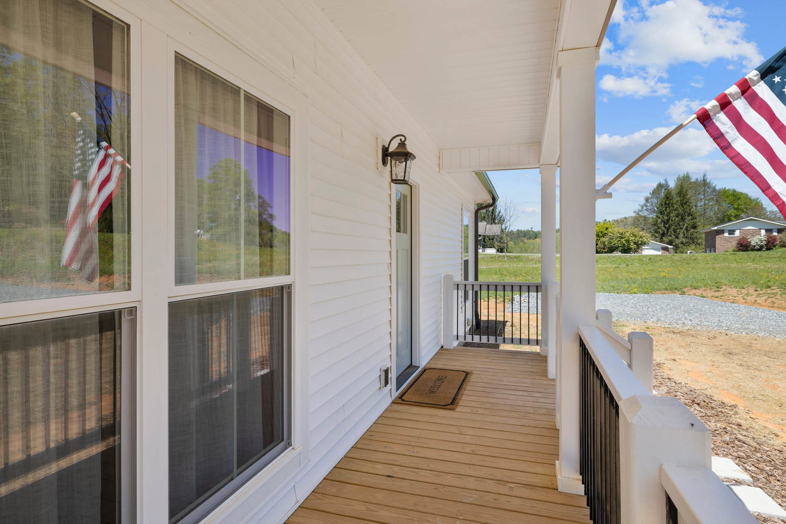 Wooden front porch with white railing, brown welcome mat featuring black text, American flag, white roof, and surrounding greenery.