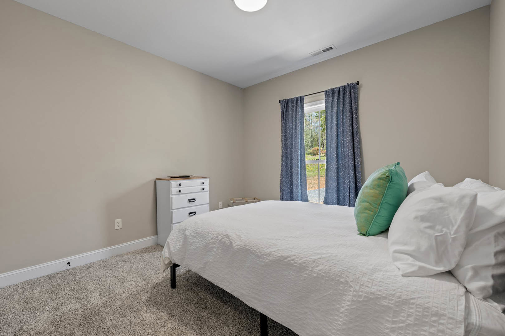 Bedroom with white bed linens, green accent pillow, white dresser with black handles, large window overlooking garden, light-colored walls, hardwood floor