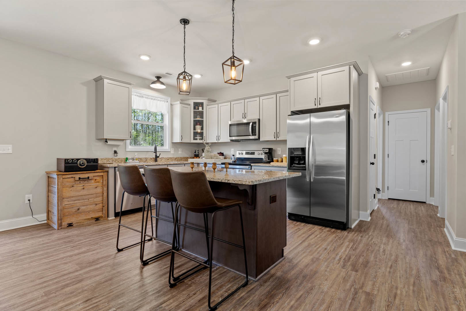 Open kitchen with white cabinetry, stainless steel refrigerator, central island topped with light stone, two wooden stools, built-in microwave, and white door with black handle.