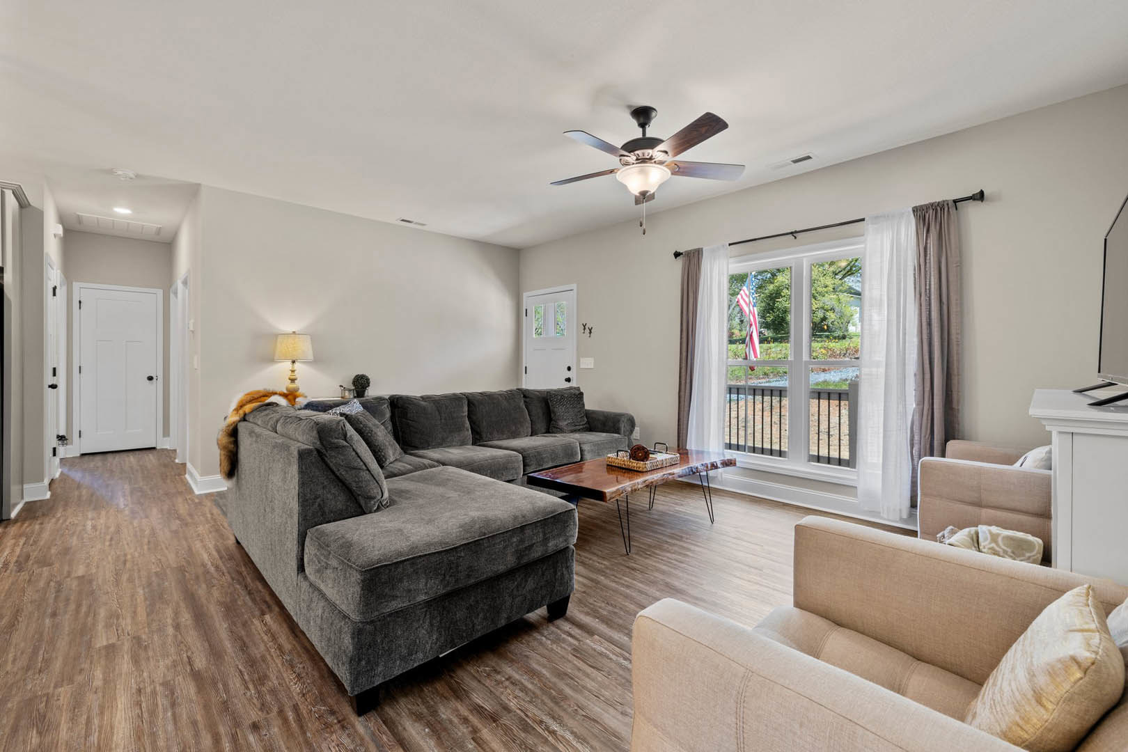 Living room with gray couch, dog resting on cushions, ceiling fan with light fixture, pillows scattered on floor, white door with black knob, window showing flag outside