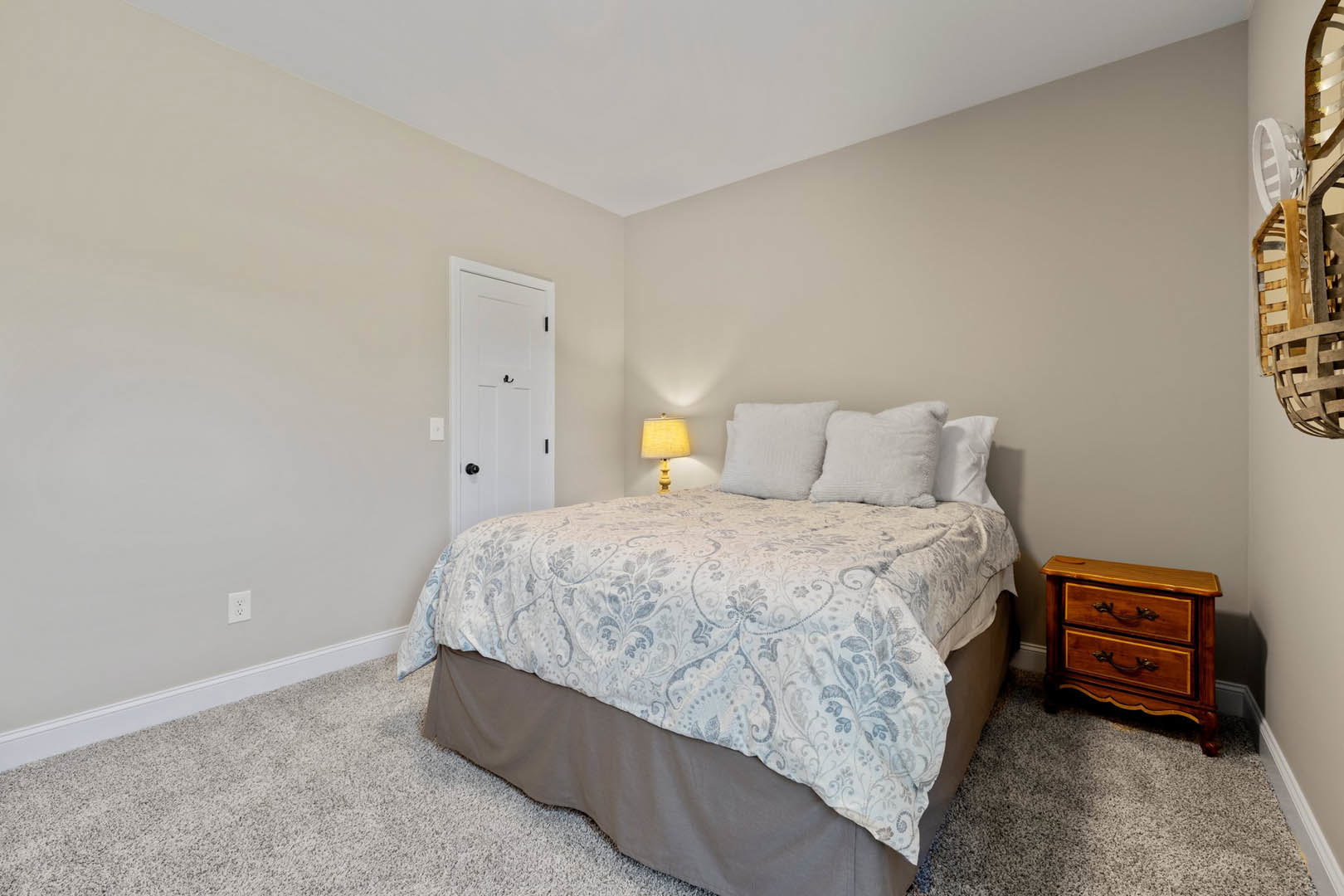 Queen bed with white bedding, yellow lamp on wood nightstand, gray dresser, white door with metal hook, neutral walls, accent pillow
