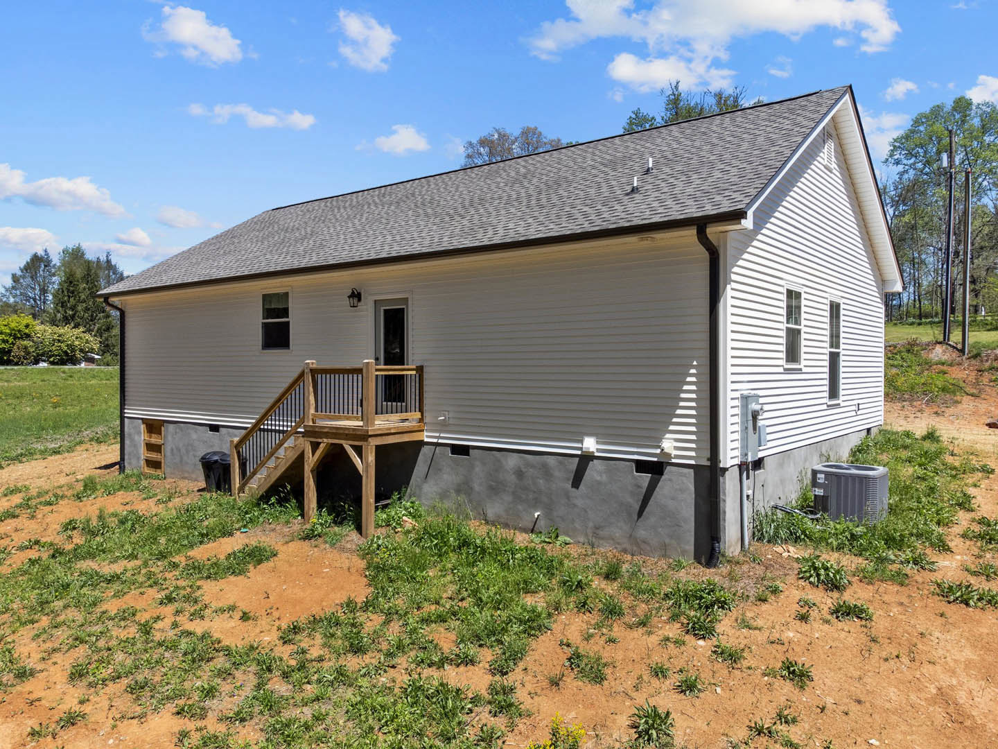 Two-story house with light siding, wooden deck and railing, wooden staircase leading to the yard, black trash can, patch of grass, and a grey rectangular object on the lawn.