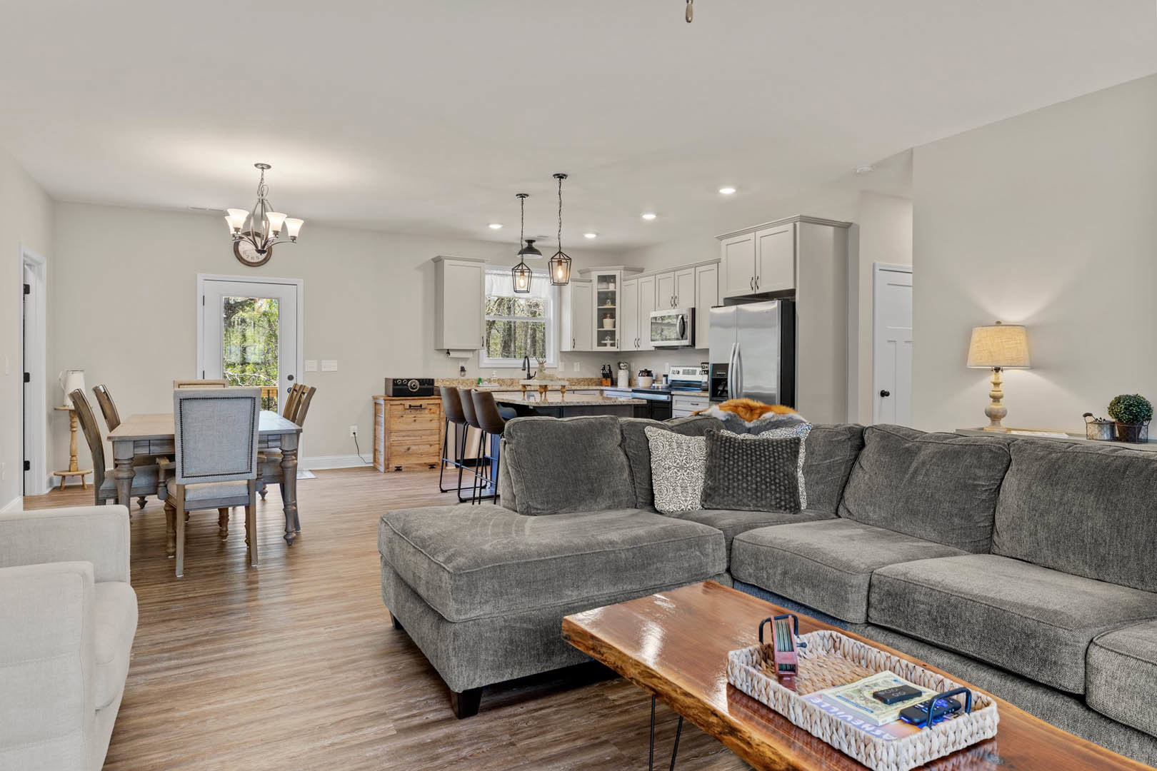 Open concept living room with a large gray sectional couch, coffee table, and sleeping cat; adjacent modern kitchen with white cabinetry; hardwood floors, neutral walls, and