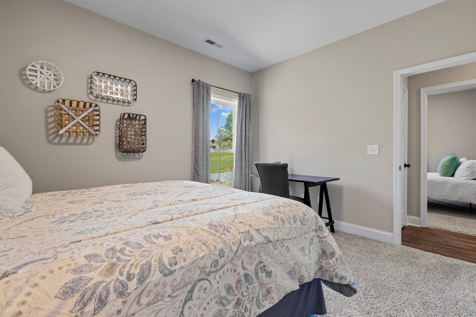 Bedroom with a bed featuring patterned sheets, a desk, pillows, and a window overlooking a grassy field; basket and white decorative object visible.