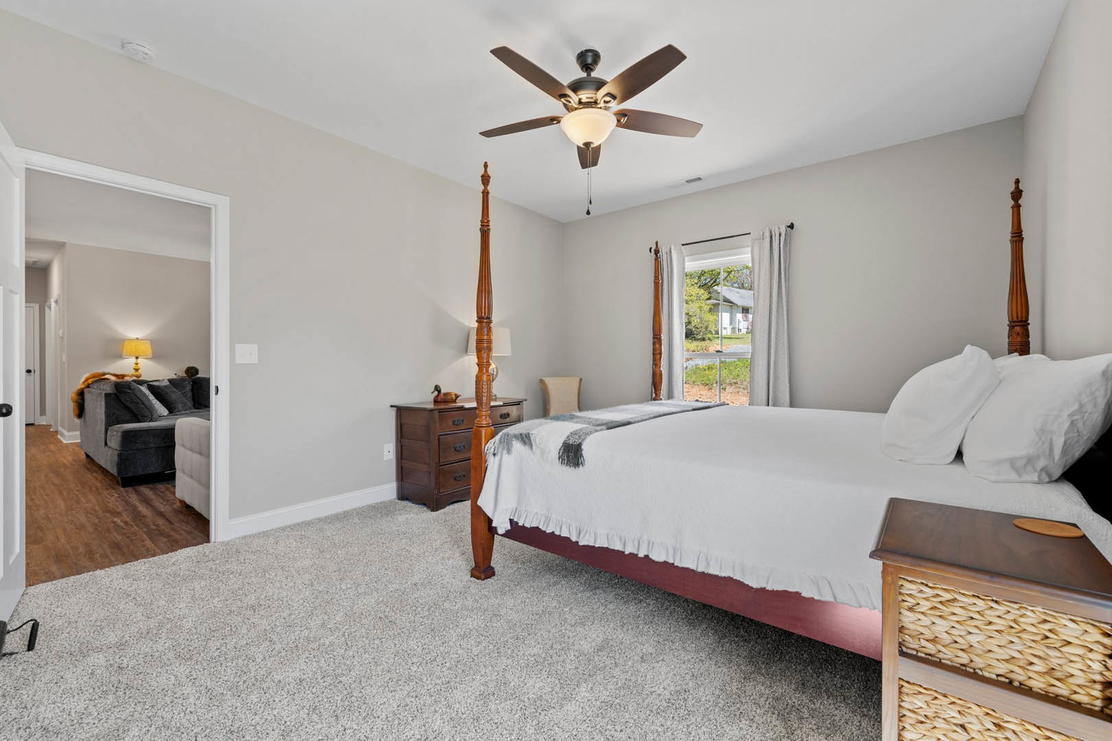 Bedroom with upholstered bed, white bedding and pillows, ceiling fan with light fixture, yellow lamp on nightstand, window showing neighboring house, couch with decorative pillows