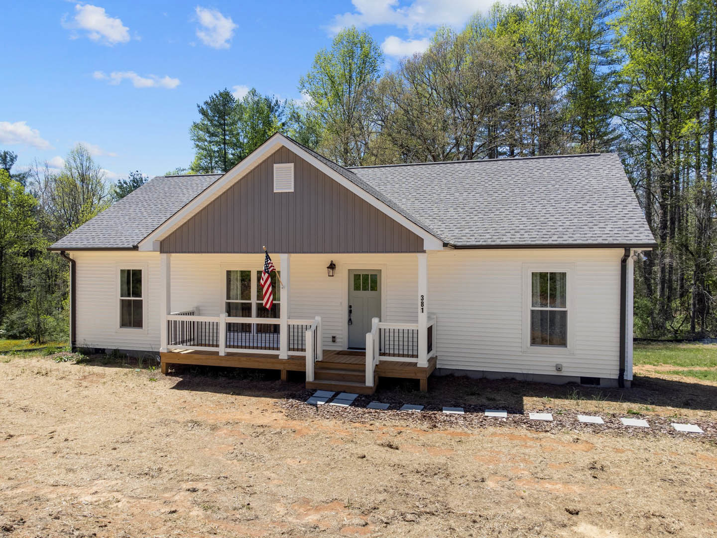 Front porch with white door and glass panes, American flag mounted near window with white frame, dirt patch and small tree in foreground, cloudy sky above light-colored cottage