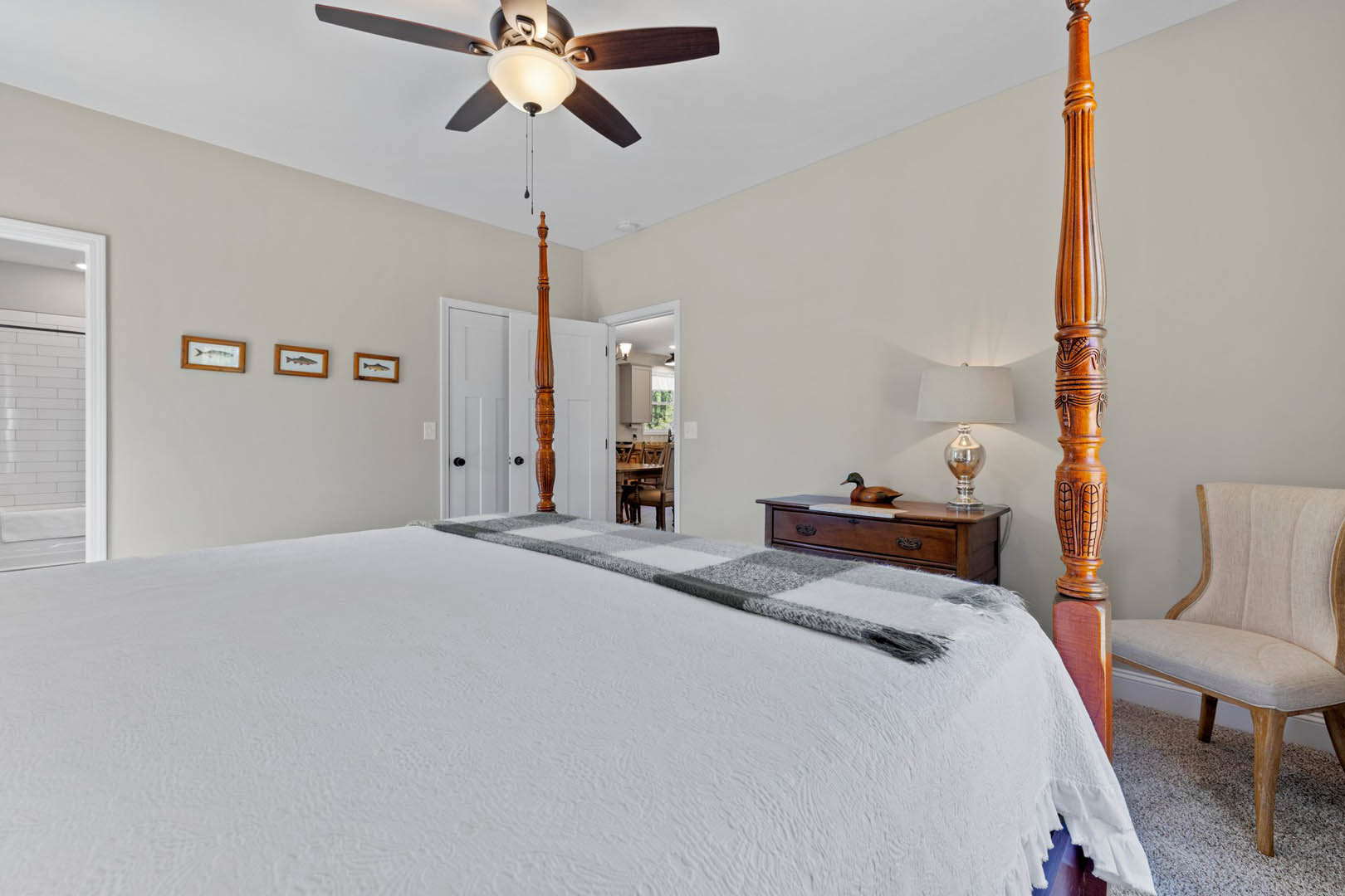 Wooden four-poster bed with white linens beneath a ceiling fan and light fixture, framed fish artwork on wall, upholstered chair, silver trophy on bedside table