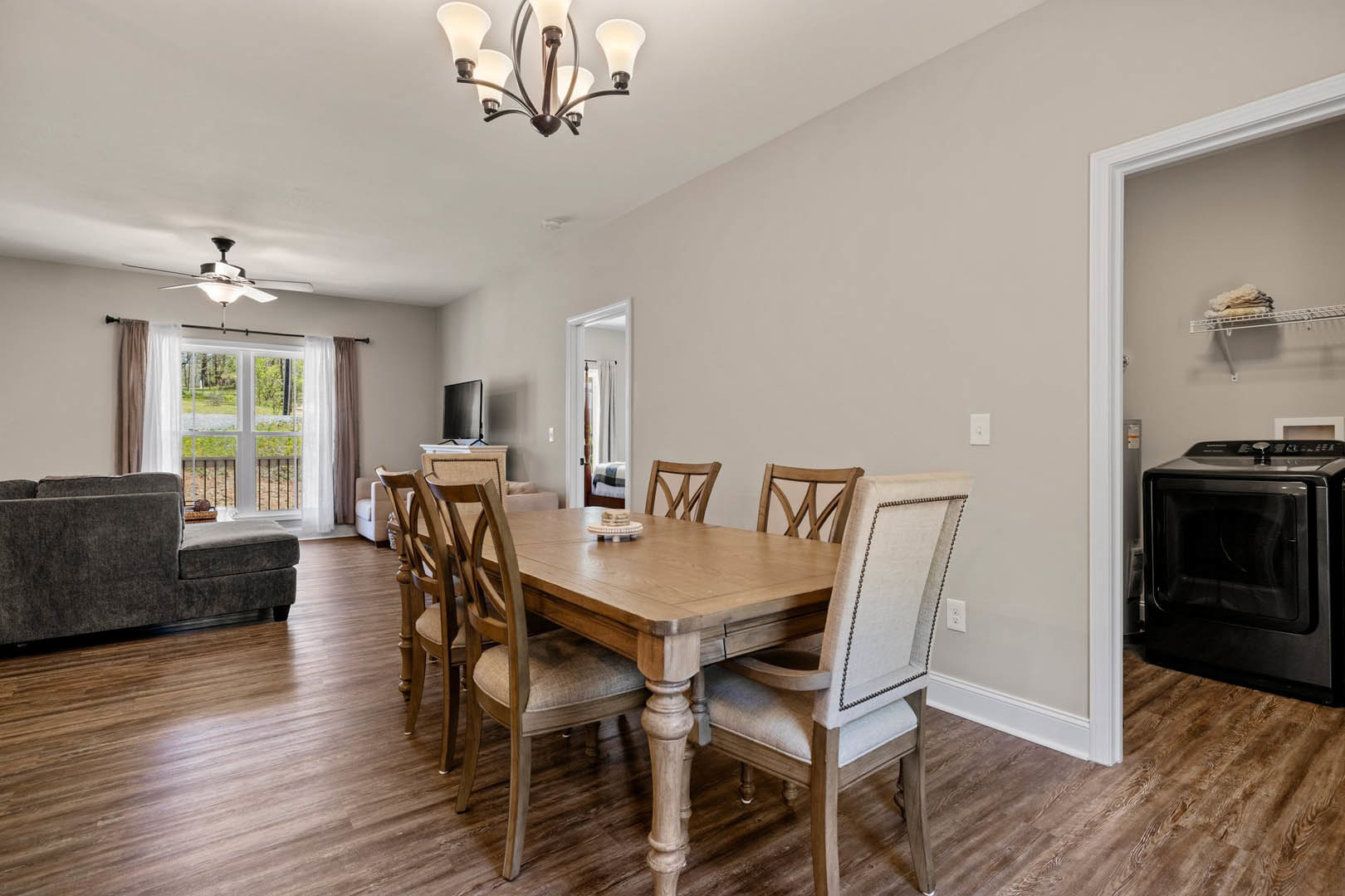Wood dining table with upholstered chairs set on hardwood floor, white pendant light fixture overhead, gray sofa in background, window with sheer curtains, black microwave on