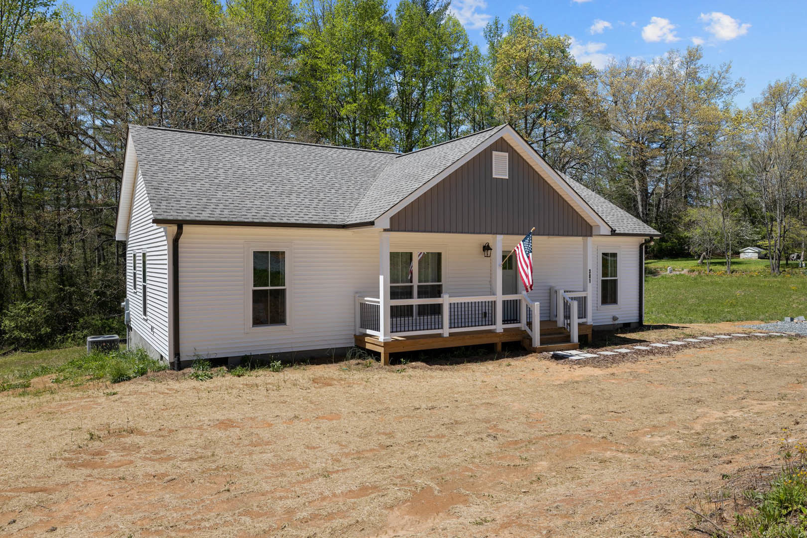 Two-story house with covered porch, white railings, American flag, brown dirt yard, large windows, surrounded by trees under blue sky with clouds