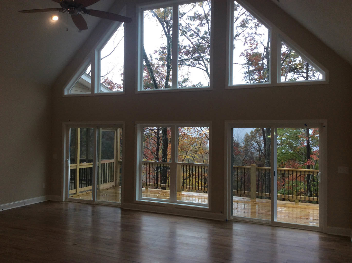 Open living room with wide plank wooden floors, large windows overlooking trees, glass door leading to deck with metal railing, ceiling fan with light fixture
