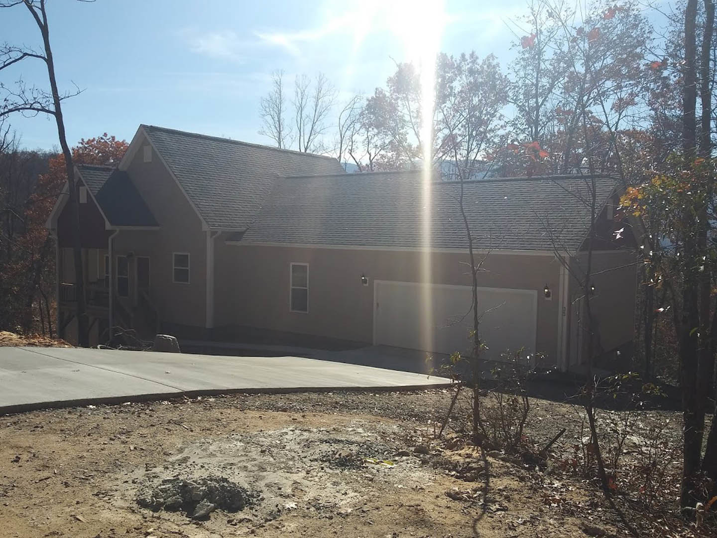 Two-story home with light siding, attached garage, concrete driveway, glass front door, mature trees and sunlight filtering through foliage in the background