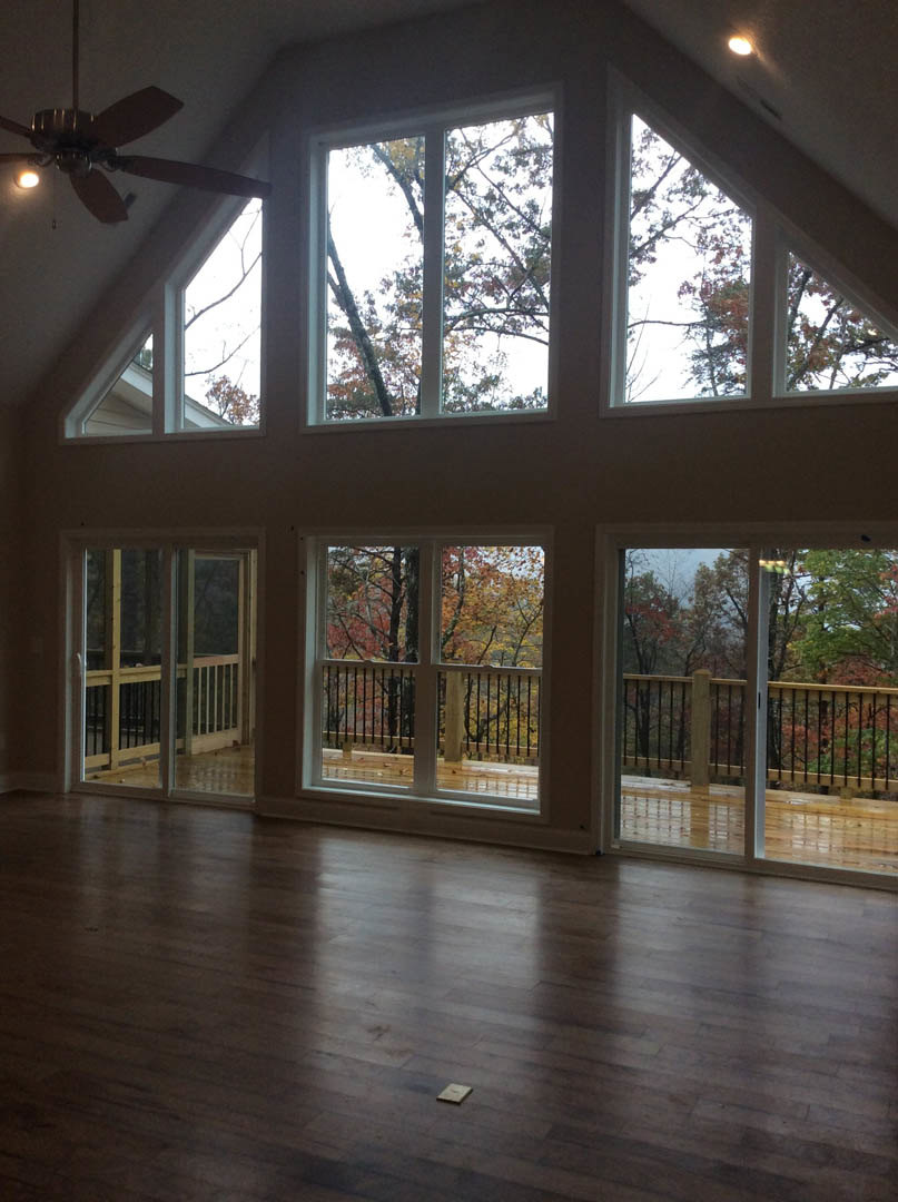 Spacious living area with wide plank wooden flooring, multiple tall windows overlooking trees, ceiling fan with light fixture, and access to a deck through glass doors