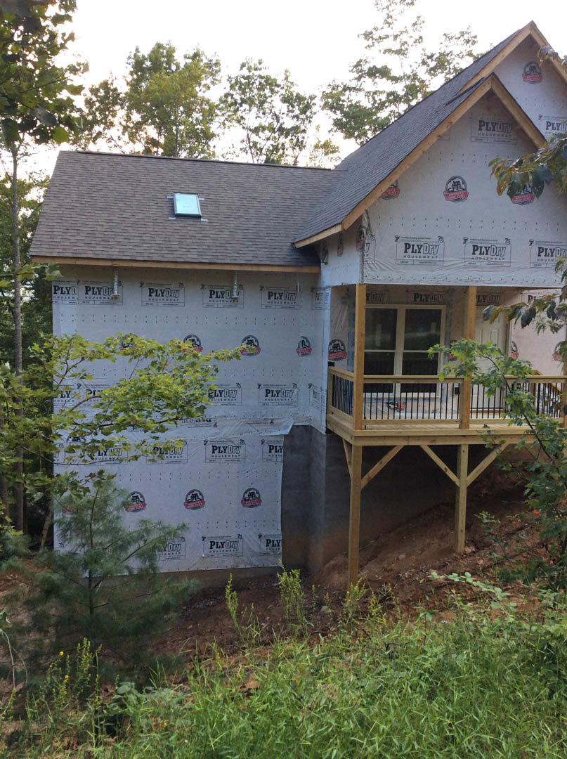 Two-story house with white siding, wooden deck, covered front porch, large windows, and mature tree in landscaped yard