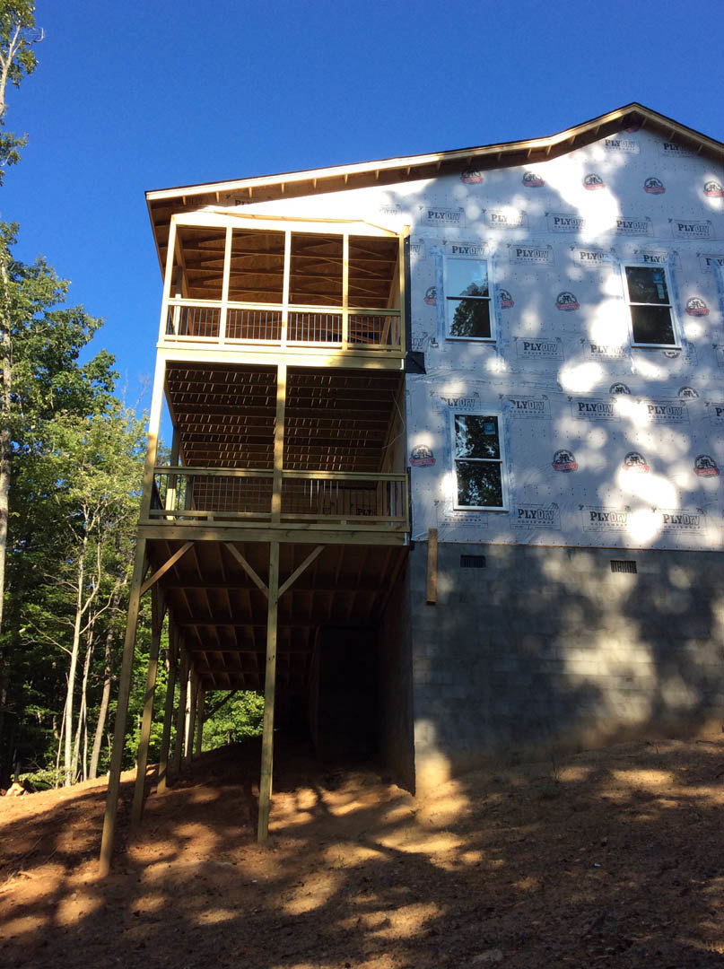 Wood-framed house under construction with exposed wooden deck, white-framed windows, dirt yard, and mature trees in the background