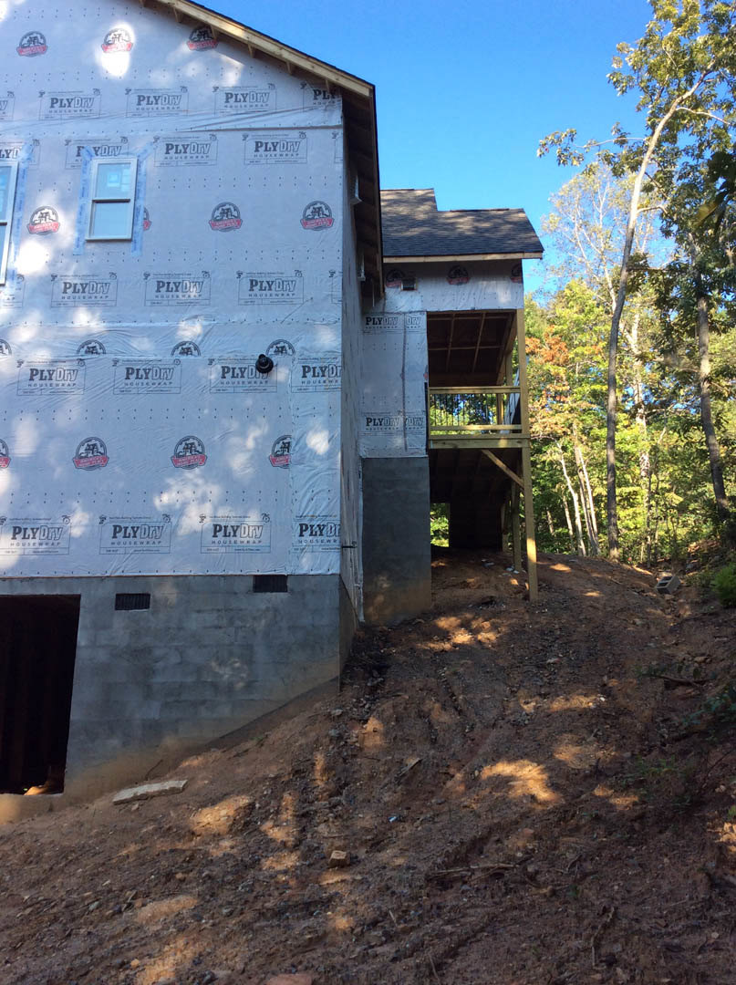 Partially built house with exposed framing, white protective sheeting, elevated wooden deck, dirt slope in foreground, leafy trees surrounding structure, blue sky overhead