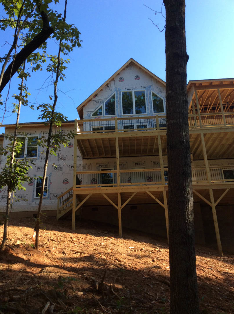 Wood deck with metal railing, tree trunk and branches in front of house, dirt ground with scattered sticks, windows featuring wooden and white frames