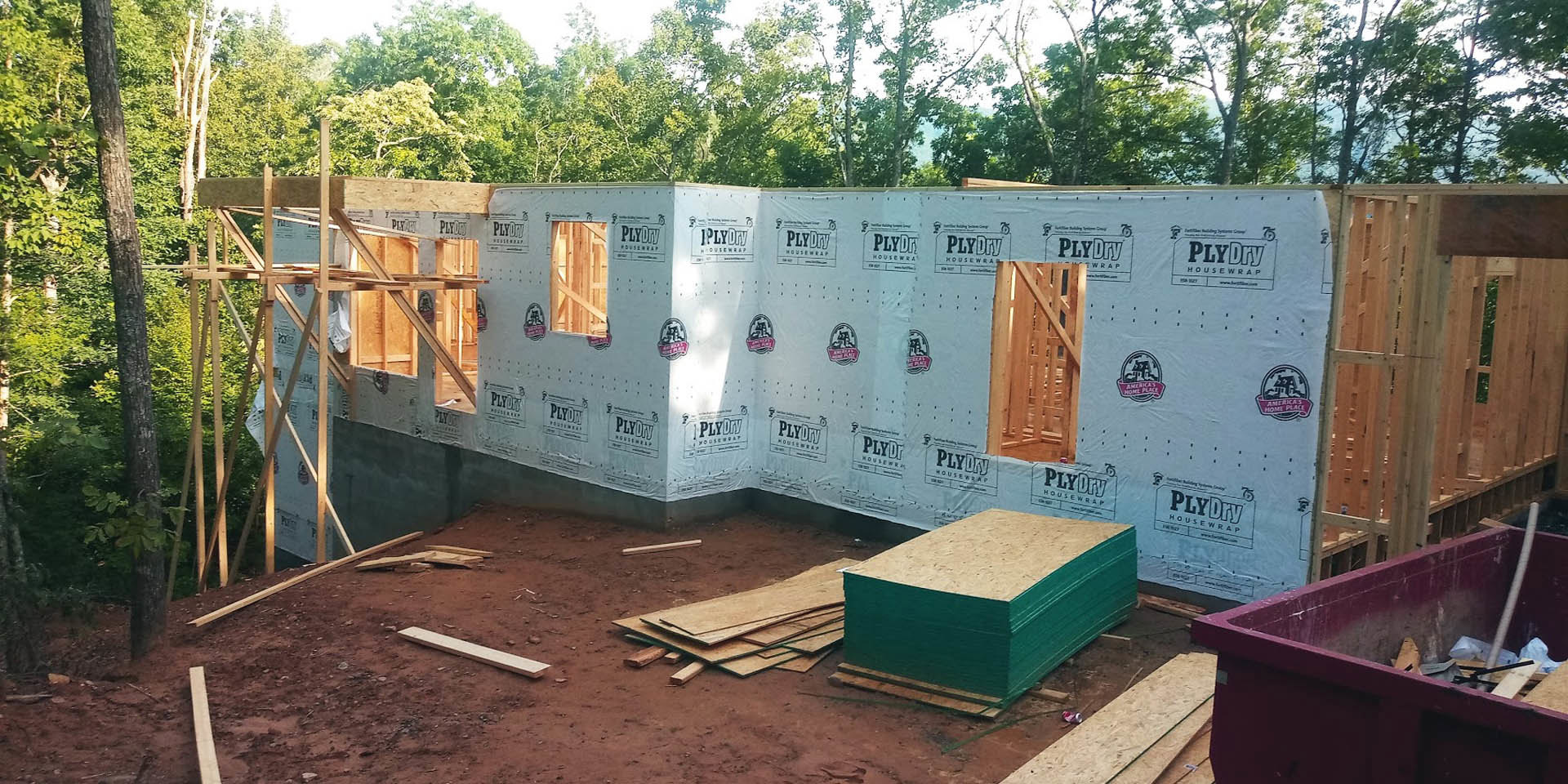 Wood-framed custom home under construction with exposed plywood sheets, partially finished exterior walls, and a wooden gate beside a blue wall; trees and mountains visible in the