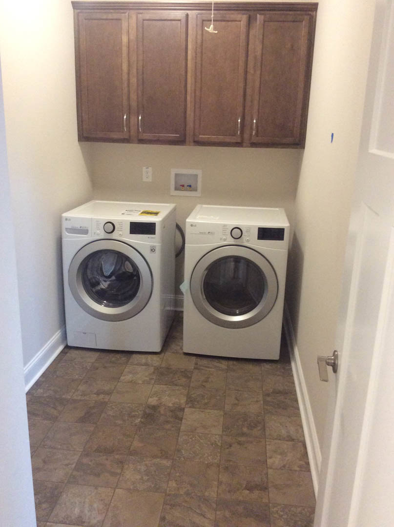 White front-loading washing machine and dryer set against white cabinets in a laundry room with brown tile flooring, colorful objects on a picture frame above appliances.