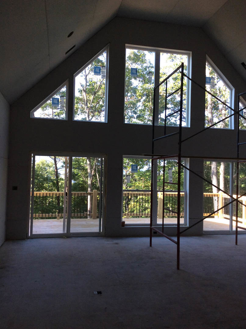 Unfinished room with concrete floor, large windows, metal scaffolding, sliding glass door opening to view of trees, and wooden deck with red railing