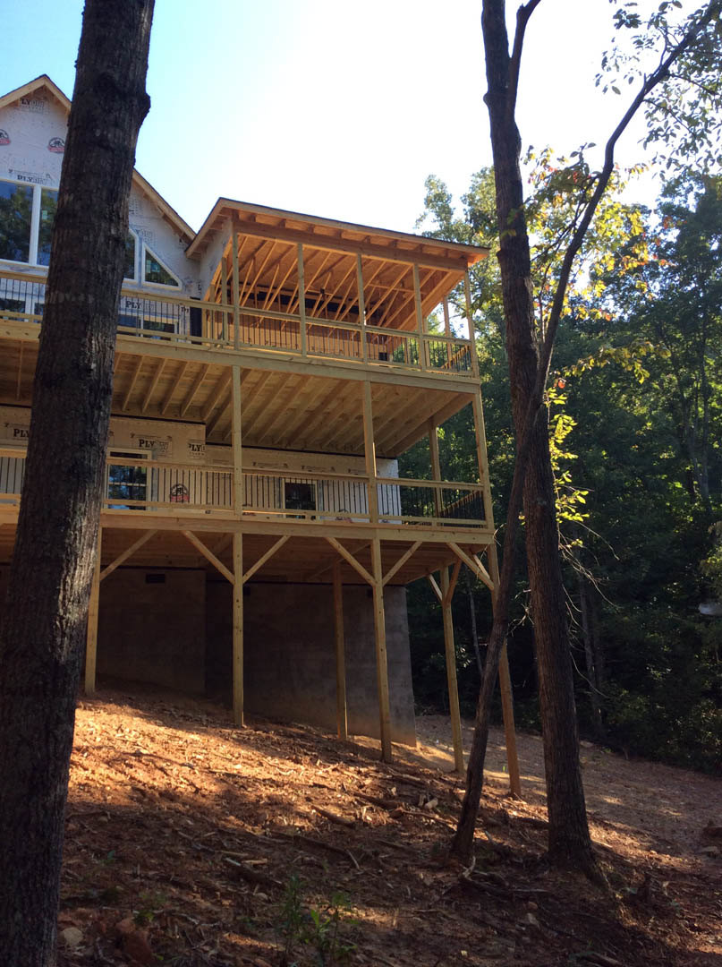 Two-story home with wood deck and covered porch, surrounded by mature trees and natural landscaping, large windows with wooden frames visible on exterior.