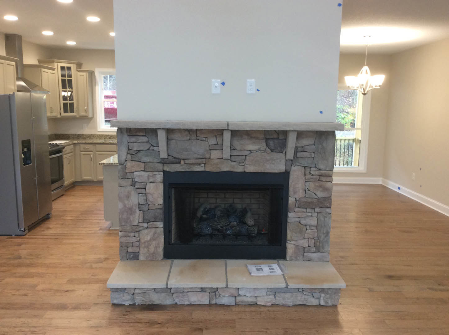Stone fireplace with stacked logs, dark metal fire screen, light wood flooring, built-in shelves, and white walls with electrical outlets