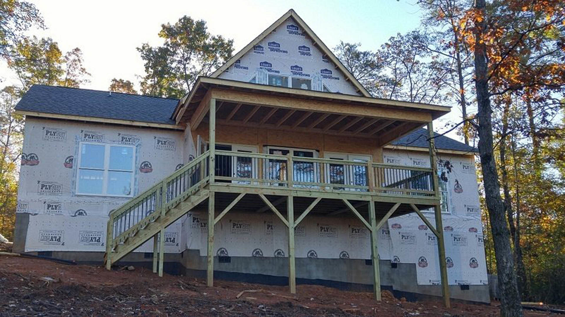 Two-story house with light siding, covered front porch, upper balcony, wooden deck with stairs, large windows, and mature tree in landscaped yard