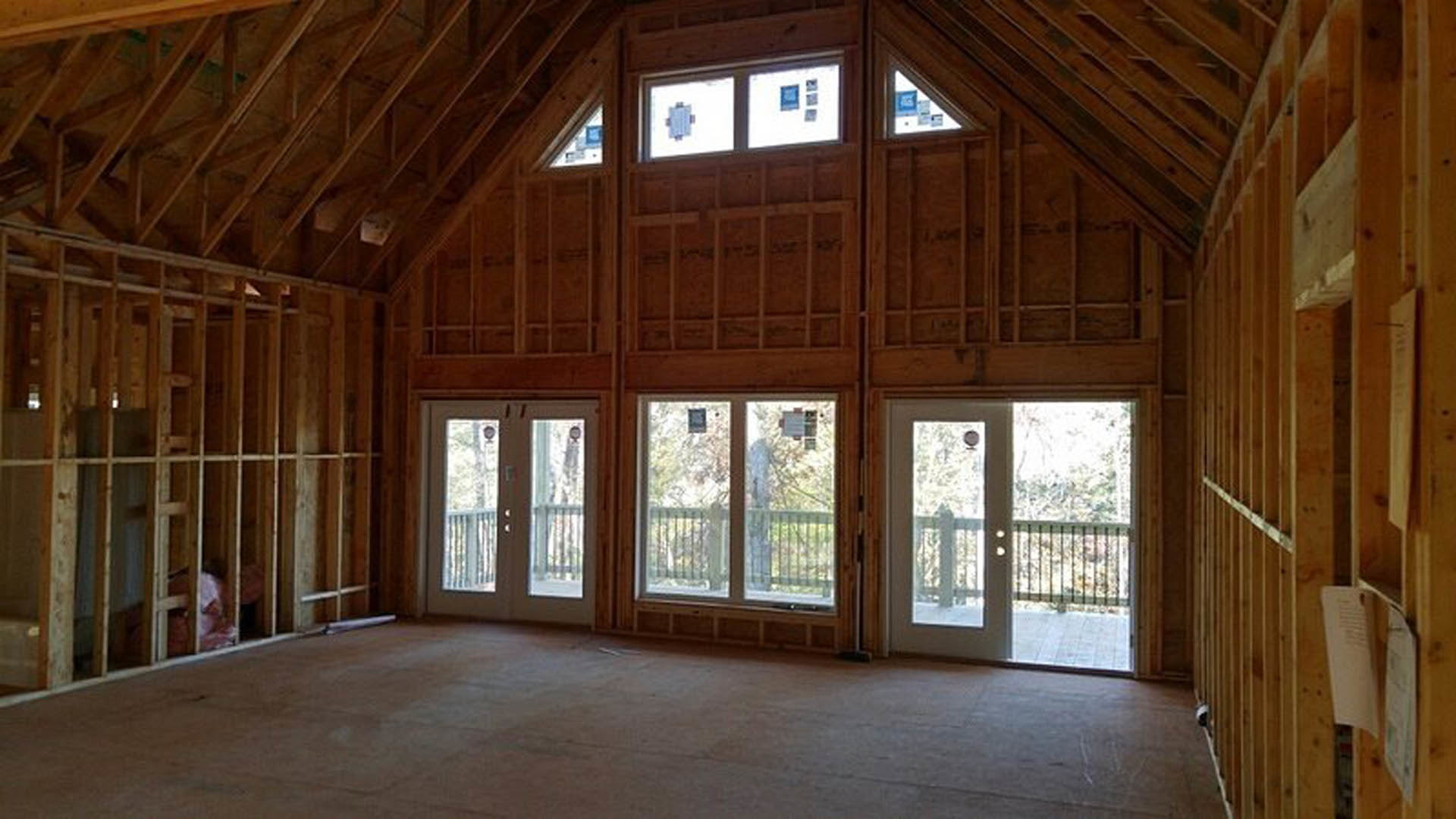 Spacious room featuring double glass doors opening to a balcony with metal railing, hardwood flooring, exposed ceiling beams, and large windows allowing natural daylight